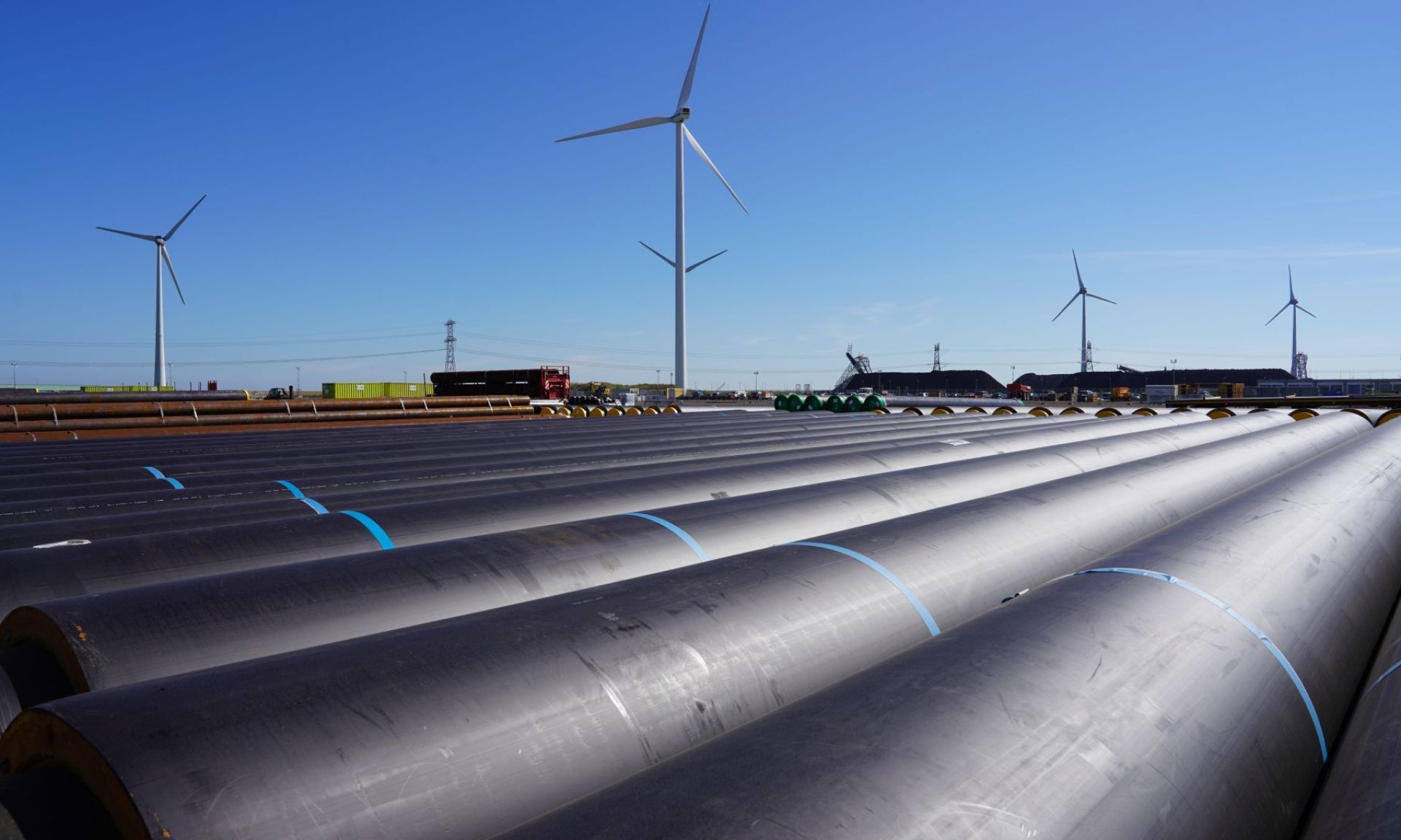 Grote pijpleidingen naast een veld met windturbines onder een heldere blauwe lucht.